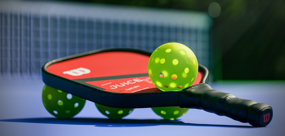 A pickleball and paddle laid on a vibrant outdoor court, bathed in natural light – a scene that captures the essentials for a game of pickleball, combining the distinct paddle and iconic perforated ball ready for play on a sunny day
