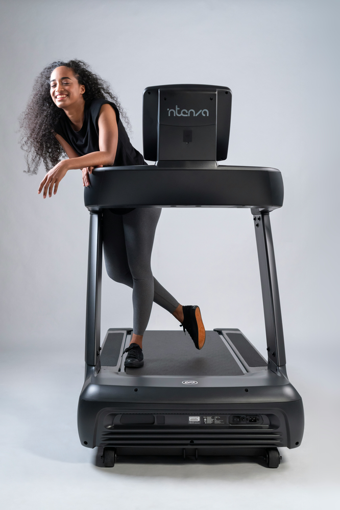a woman standing confidently next to a treadmill, ready for a workout. She is wearing workout attire and exudes a positive and determined demeanor, suggesting an active and healthy lifestyle.