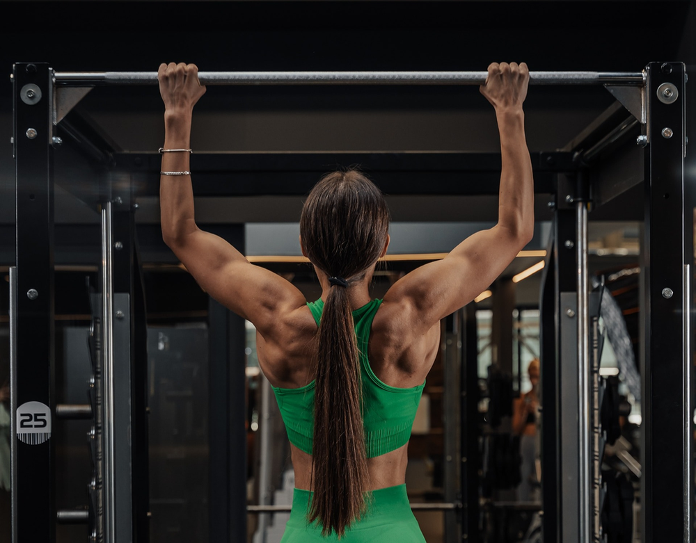 Woman performing pull-ups, showcasing upper body strength and fitness. Engaged in bodyweight exercise, highlighting determination and active lifestyle. Gym environment with pull-up bar in the background, emphasizing commitment to strength training