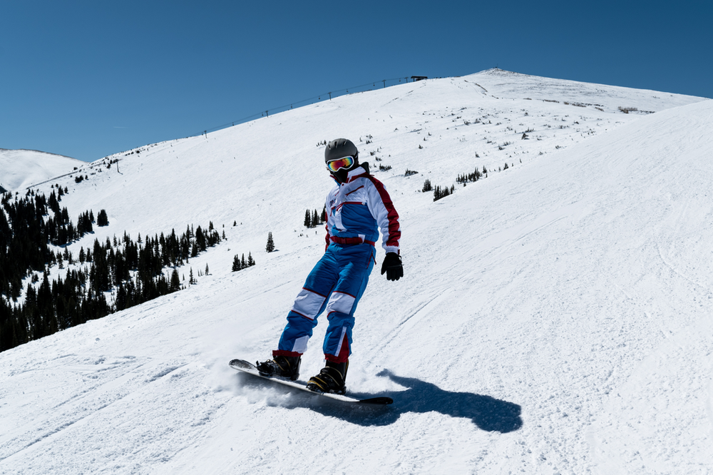 A snowboarder carving through fresh powder on a snow-covered mountain slope, leaving a trail of spray behind. The rider is dressed in winter gear, confidently navigating the snowy terrain with a snow-capped mountain landscape in the background.